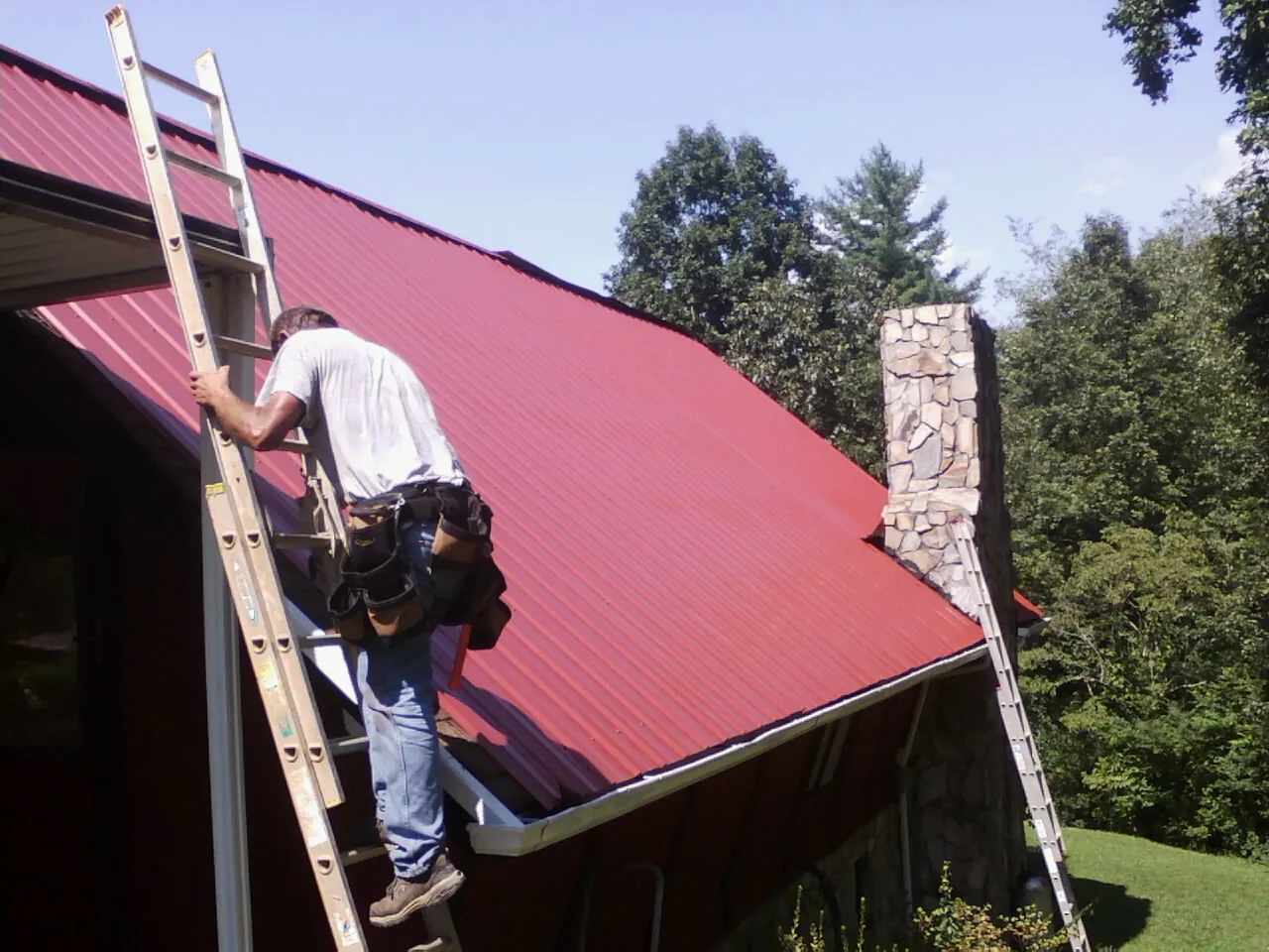Roofer on a ladder beside a red metal roof and stone chimney — Soffit & Fascia Repair by Roofing Brothers in Flag Pond